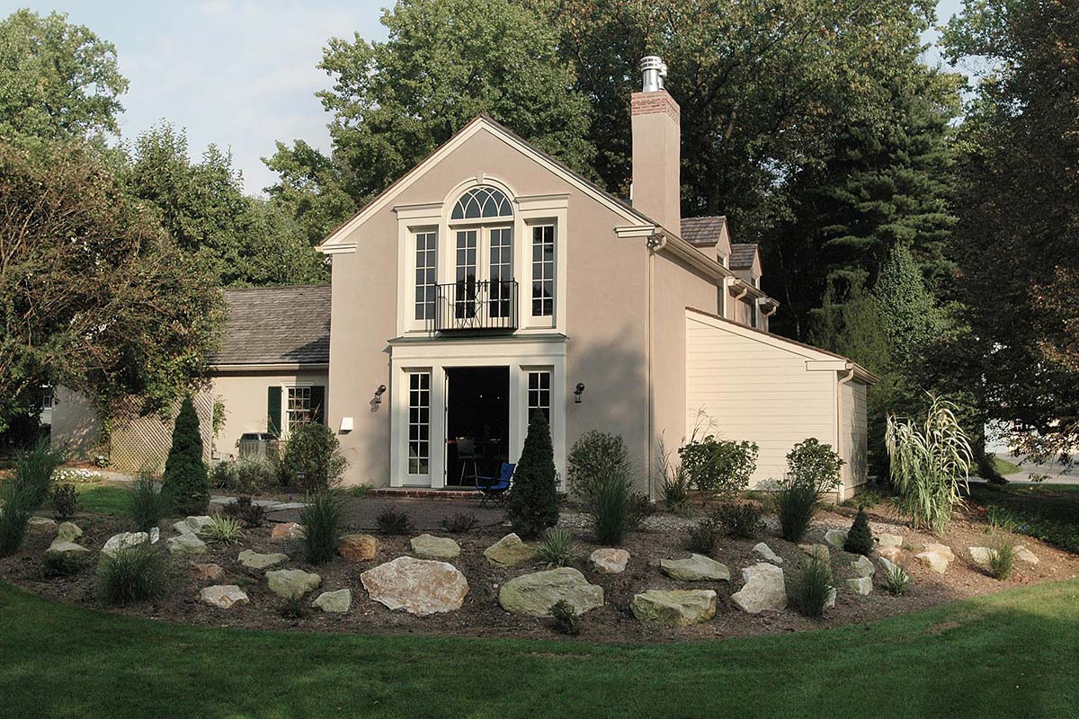 Elegant home with arched balcony and large windows, surrounded by lush greenery and a rock garden.