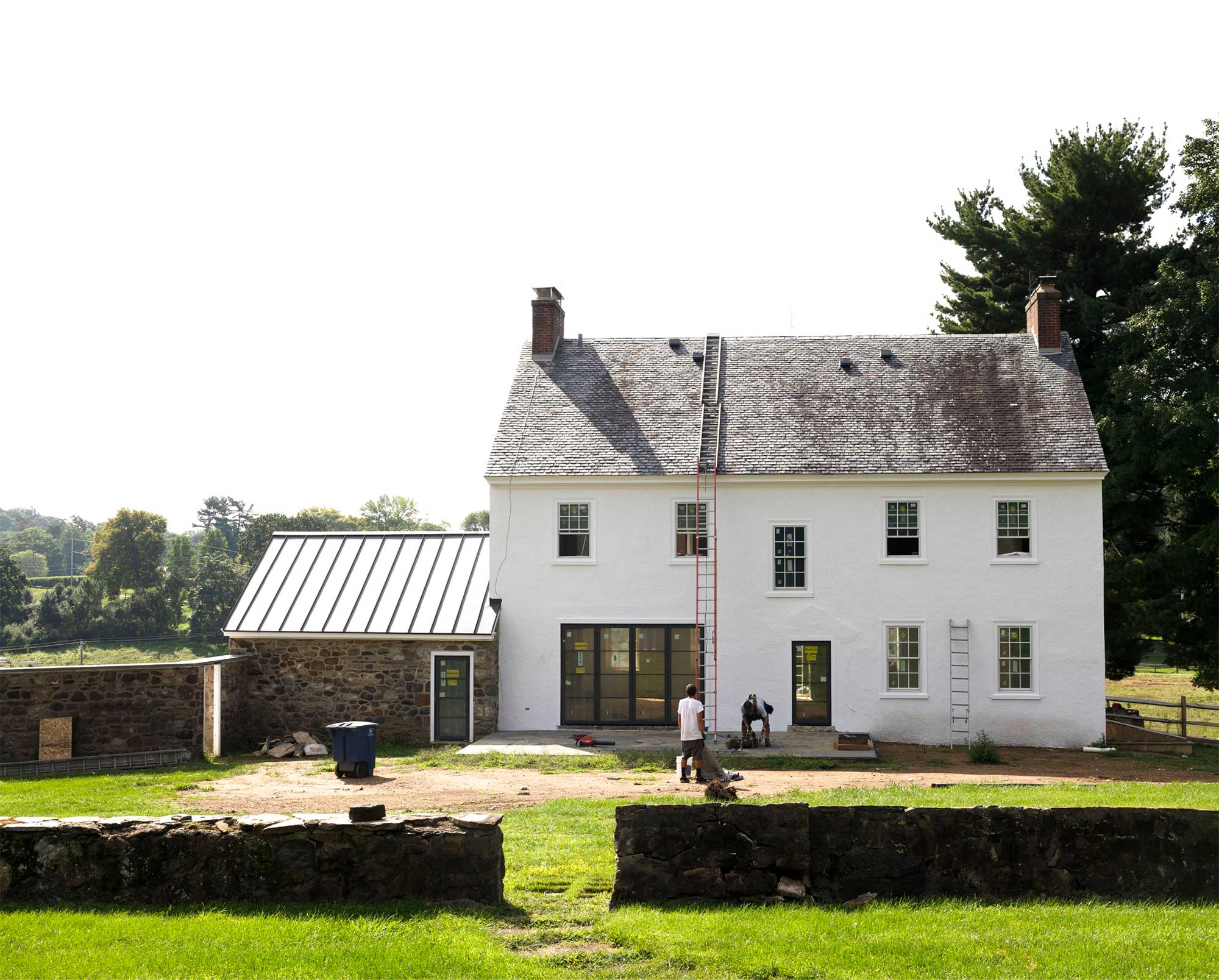 Two workers with ladders in front of a white historic home with stone facade and metal roof, surrounded by stone walls and greenery.
