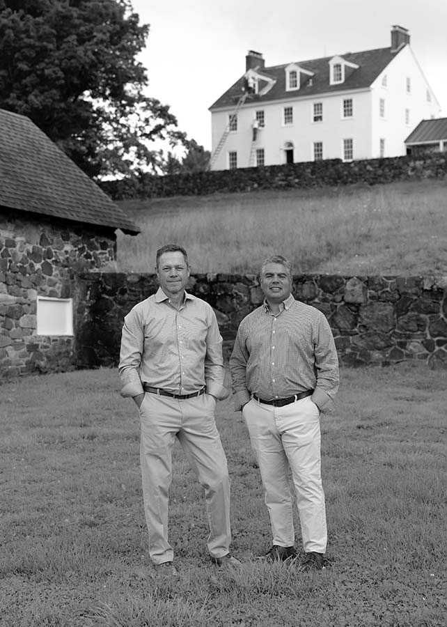 Black and white photo of Andy Kerns and Greg Ranieri, Ranieri & Kerns founders, standing before a historic house with stone walls and lush greenery.